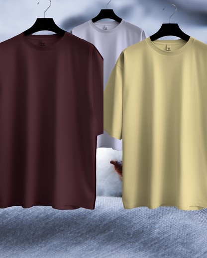 Three plain t-shirts in brown, white, and beige on hangers against a textured gray background.
