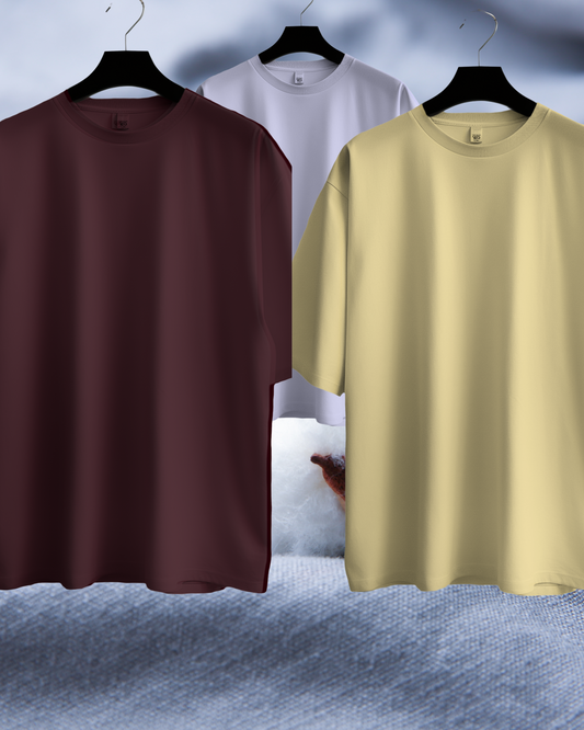 Three plain t-shirts in brown, white, and beige on hangers against a textured gray background.