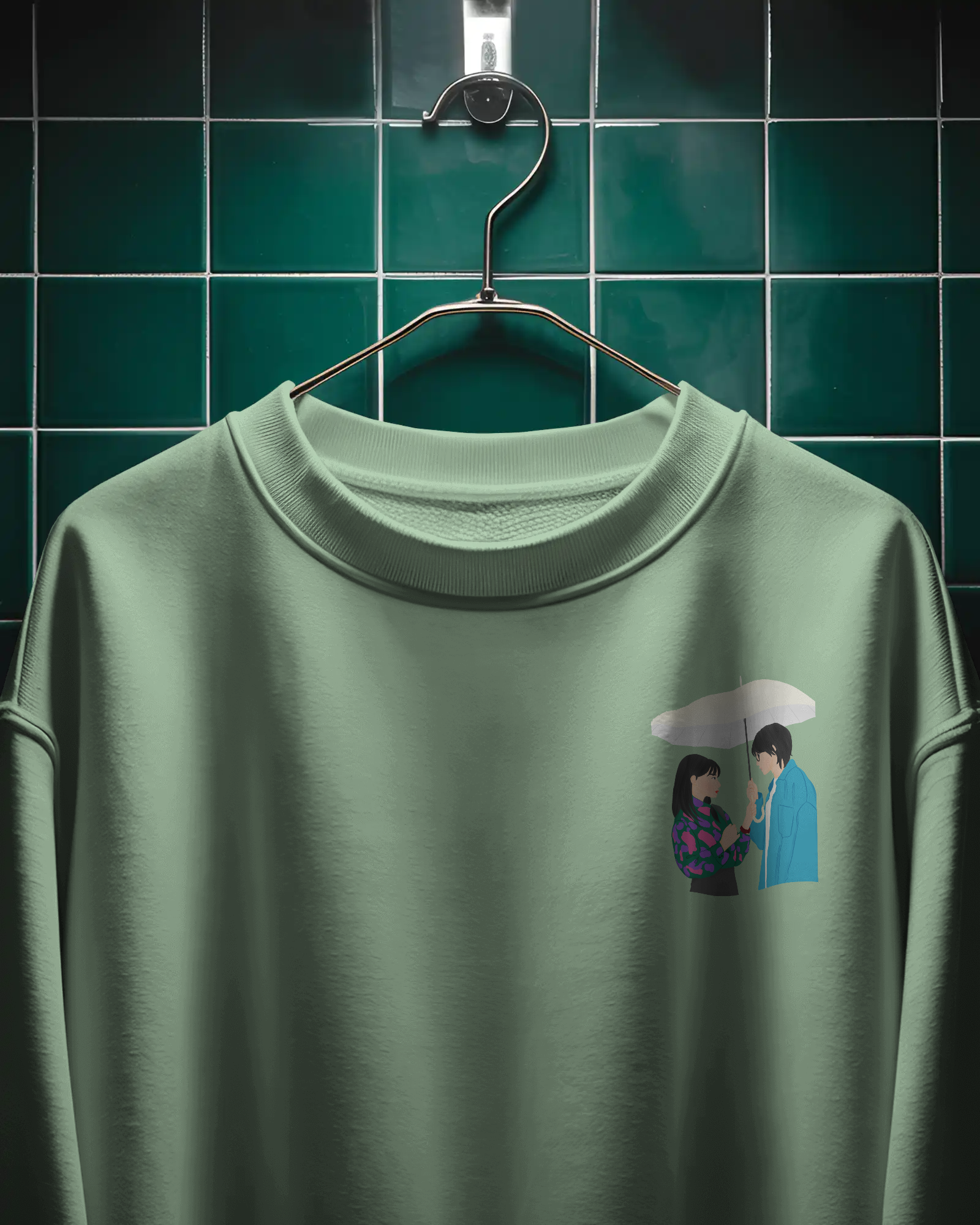 Green sweatshirt with a graphic of two people under an umbrella on a hanger against a green tiled wall.