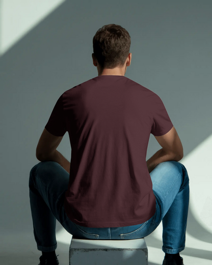Man wearing a maroon t-shirt and blue jeans sitting on a white stool against a gray wall.